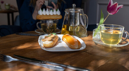 Enjoy a serene afternoon tea setting with a variety of pastries, including eclairs, served on a wooden table bathed in sunlight. The ambiance is complemented by a vibrant tulip