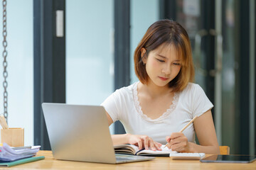Charming asian businesswoman working on laptop and taking note while sitting at the table in office.