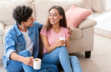 Black teen couple sits together on the floor of their home, enjoying coffee and conversation. The girl smiles as she looks at the guy, who is holding her arm. The couple is dressed in casual clothes