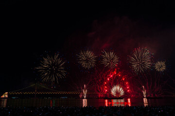 a nighttime fireworks display over water in Busan, Korea, with a crowd of spectators and a bridge illuminated in the background.