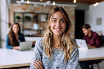 Fototapeta premium A friendly, smiling woman poses with her arms crossed in a contemporary office setting, with colleagues soft in focus behind