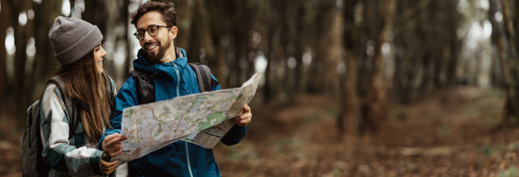 A couple hikes through a forest while looking at a paper map. The man is smiling and holding the map, while the woman is looking at him and smiling, panorama with copy space