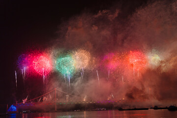 a vibrant fireworks display over a bridge at night, reflecting on water with spectators in silhouette. It’s a beautiful scene in Busan, Korea.