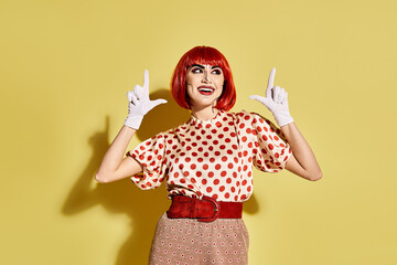 A pretty redhead woman with pop art makeup wearing a polka dot blouse and white gloves against a yellow background.
