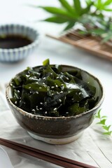 A bowl of fresh seaweed salad topped with sesame seeds, accompanied by a dipping sauce and chopsticks on a white cloth.