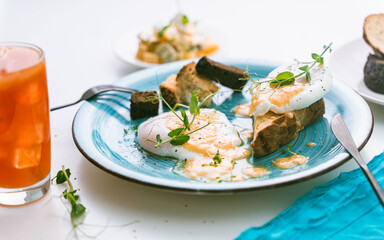 Eggs Benedict with fried toast and crouton on a turquoise plate and orange juice with ice on a white background. Selected focus
