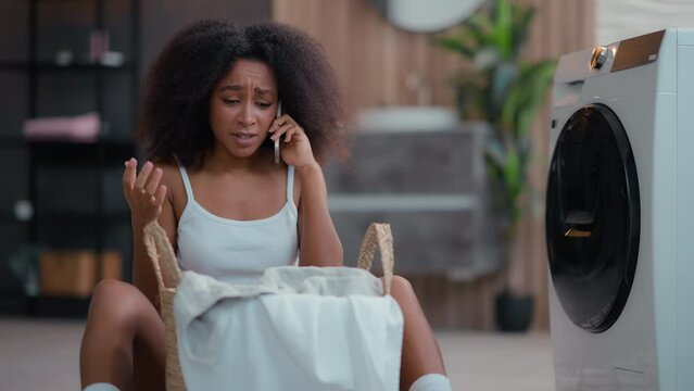Upset woman housewife African American frustrated female stressed girl with laundry basket dirty clothes talking negative mobile phone call washing machine broken calling repair service talk cellphone