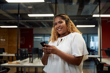 Young multiracial woman in businesswear smiling while using mobile phone in office