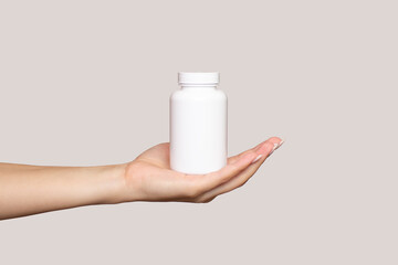 The hand of a young woman holds a white plastic bottle on her palm, isolated on a beige background....