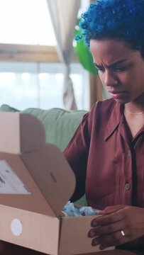 Young frustrated ethnic African American woman shopaholic gets upset after unboxing parcels from online store due to low quality or wrong size of products sits on sofa in apartment.