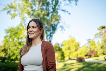 Smiling woman in a white top and brown cardigan, standing in a lush green park on a sunny day, looking to the side with trees in the background. 