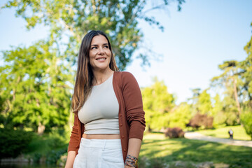 Woman in a white top, brown cardigan, and white pants, smiling and standing in a lush green park on a sunny day, looking to the side. 