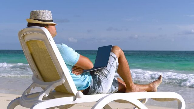 SLOW MOTION. Young man with laptop on beach sleeping on his deck chair. Man sunbathing on beach. Rest while freelancer work on beach. Caucasian man resting on sun bed by sea, holiday travel vacation