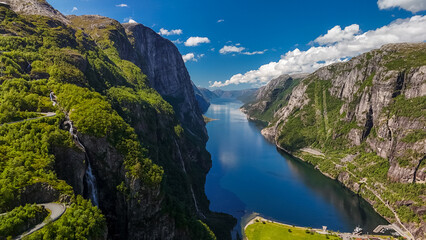 An aerial view of a picturesque fjord in Norway, showcasing towering cliffs, lush greenery, a winding road, and a stunning blue body of water. Kjerag, Lysebotn, Lysefjorden, Norway
