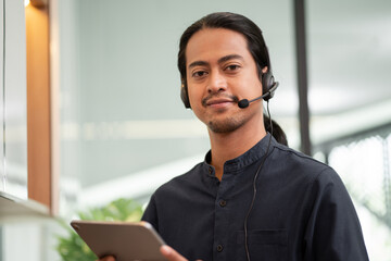 Asian male call center with digital tablet, customer service, telesales wearing headset or headphone talking with customer looking at camera