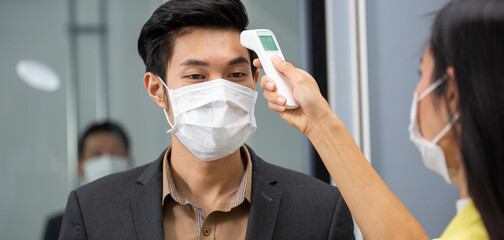 Asian employee woman using infrared thermometer check fever temperature measurement of office worker wearing face mask before enter room at entrance door