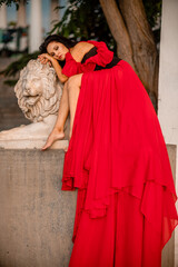 A woman in a red dress is sitting on a stone lion statue. The statue is white and has a lion's head. The woman is wearing a black belt and is looking at the camera. The scene has a calm.