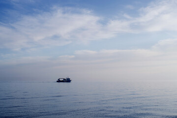 Naklejka premium Fishing boat on the horizon against the background of a blue calm sea and a cloudy sky on a summer evening