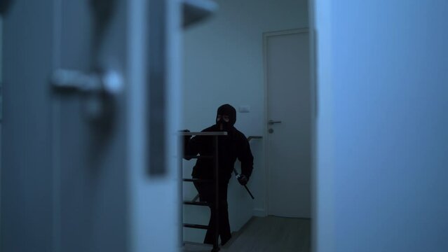 The thief with black mask holding crowbar and looking around for robbery while walking inside the apartment, home security to prevent the crimination, criminal impact on economic society.
