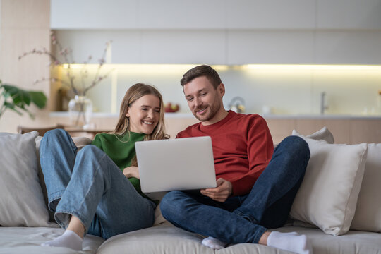 Joyful married couple watching fun movie using laptop, smiling sitting on comfortable couch in living room. Relaxed happy man and woman looking on screen computer laughing, enjoy lazy time together.