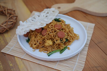 A plate of seasoned instant noodles is garnished with sliced sausage, leafy greens, and a piece of crispy cracker.