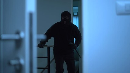The creepy burglar with black mask holding crowbar and looking around for robbery then entering inside the apartment, home security to prevent the crimination, criminal impact on economic society.
