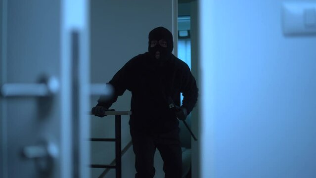 The creepy burglar with black mask holding crowbar and looking around for robbery then entering inside the apartment, home security to prevent the crimination, criminal impact on economic society.
