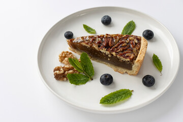 Pecan pie on a plate. Blueberries, nuts and mint leaves for dessert decoration. A yellow plate on a white background close-up.