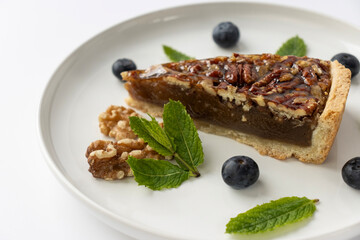 Pecan pie on a plate. Blueberries, nuts and mint leaves for dessert decoration. A yellow plate on a white background close-up.