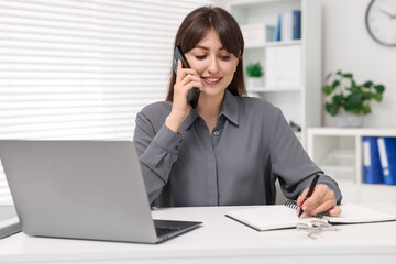 Smiling secretary talking by smartphone at table in office