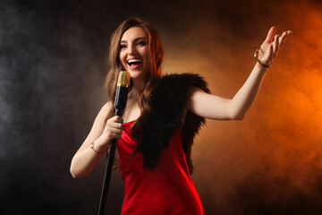Beautiful young woman in stylish dress with microphone singing on dark background in color lights and smoke