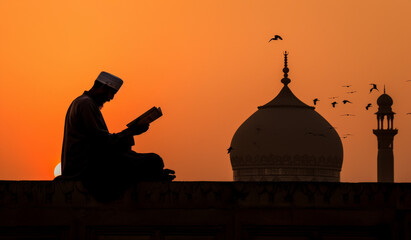 Obraz premium A muslim male figure’s silhouette is set against the orange sky of dusk, near a mosque, reading quran
