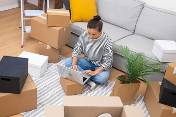 Woman in freshly acquired apartment, amidst boxes, works on her laptop, exploring interior design,...