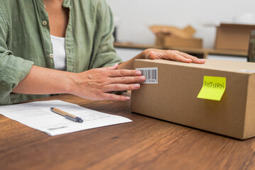 Hands of female consumer stick barcode on cardboard box with yellow sticker with word Return sitting at workplace with statement concept of returning parcel