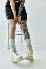 A young woman with tattoos on her leg and arm wears a plaid skirt and white boots.