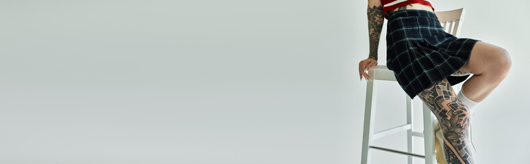 A young woman with tattoos on her legs, wearing a plaid skirt and tank top, sits on a stool against...