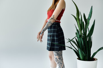 A young woman with tattoos on her arms and legs poses against a grey wall, wearing a plaid skirt and a red tank top.