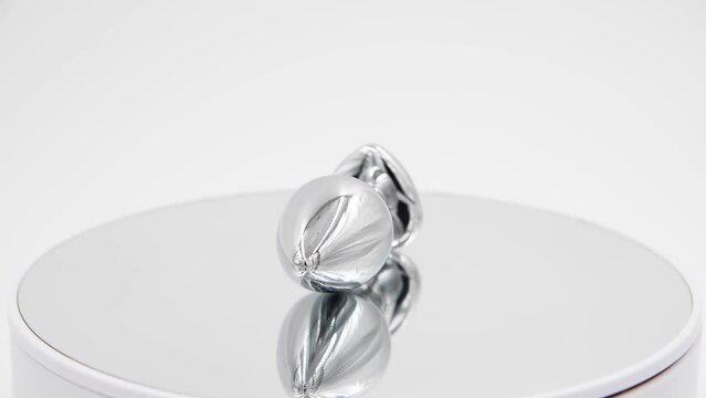 Close-up side view of metallic butt plug with pink heart shaped crystal spinning on mirror surface against white background. Soft focus. Real time video. Adult sex toys theme.	