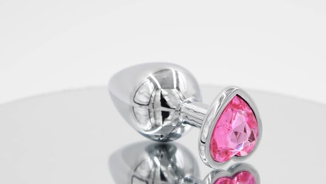 Close-up side view of metallic butt plug with pink heart shaped crystal spinning on mirror surface against white background. Soft focus. Real time video. Adult sex toys theme.	