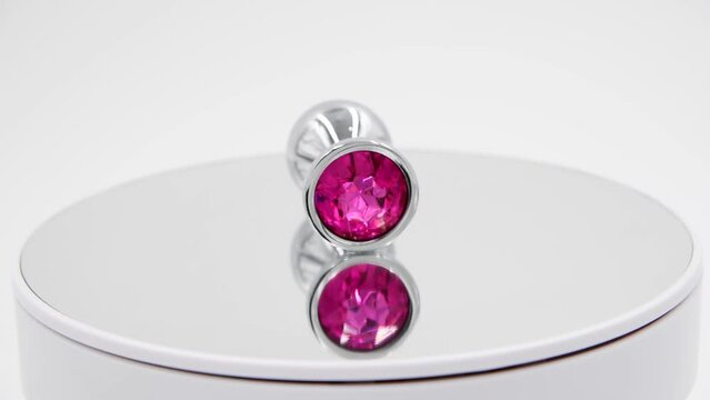 Close-up side view of metallic butt plug with pink round shaped crystal spinning on mirror surface against white background. Soft focus. Real time video. Adult sex toys theme.
