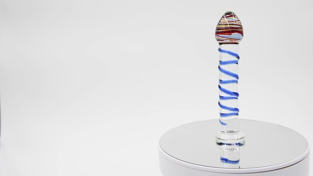 Close-up side view of bright glass dildo with various lines spinning on mirror surface against grey background. Soft focus. Real time video. Sex shop display. Copy space. Adult sex toys theme.