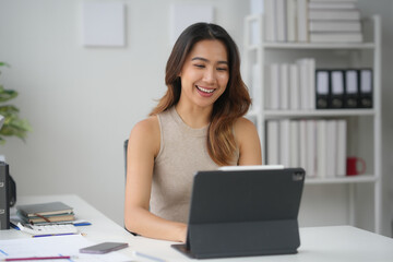 Smiling woman working on a tablet at a modern office desk, surrounded by books, papers, and office supplies.