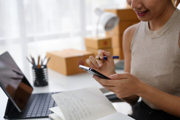 Woman using a smartphone while taking notes in a notepad, working from home with a laptop and shipping boxes in the background.