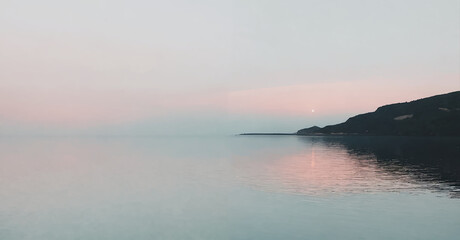 Fototapeta premium A pink and gray sunset over a calm sea with a dark shoreline in the foreground.