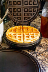 A finished waffle sits on the waffle iron with batter cup and honey in background.