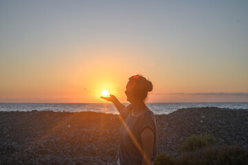 Silhouette of a young Caucasian woman holding the sun in her hands. On the seashore at sunset.