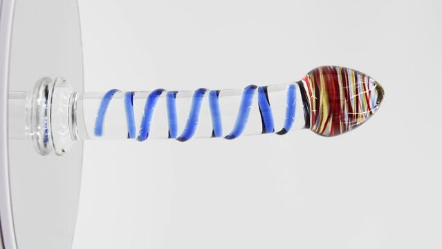 Close-up side view of bright glass dildo with various lines spinning on mirror surface against gray background. Soft focus. Real time vertical video. Sex shop display. Adult sex toys theme.