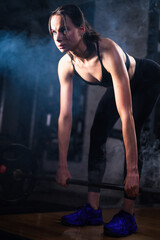 Medium shot of a female young athlete exercising deadlift in a misty dark gym with moody environment