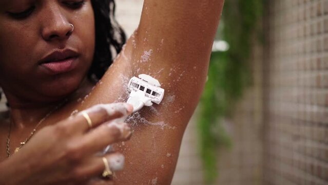 Young black woman shaving her armpits in the shower. Latin female removing her hair with a soft razor in the bathroom of her home. Body care routine and skin cosmetics. Copy space video.