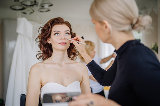 Valmiera, Latvia - September 9, 2023 - Bride getting her makeup done by a makeup artist, preparing for the wedding, with her wedding gown hanging in the background...